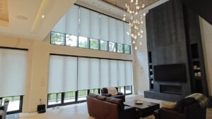 Modern living room with large floor-to-ceiling white roller blinds on expansive windows, featuring a crystal chandelier, leather sofa, and dark stone fireplace in a double-height space.